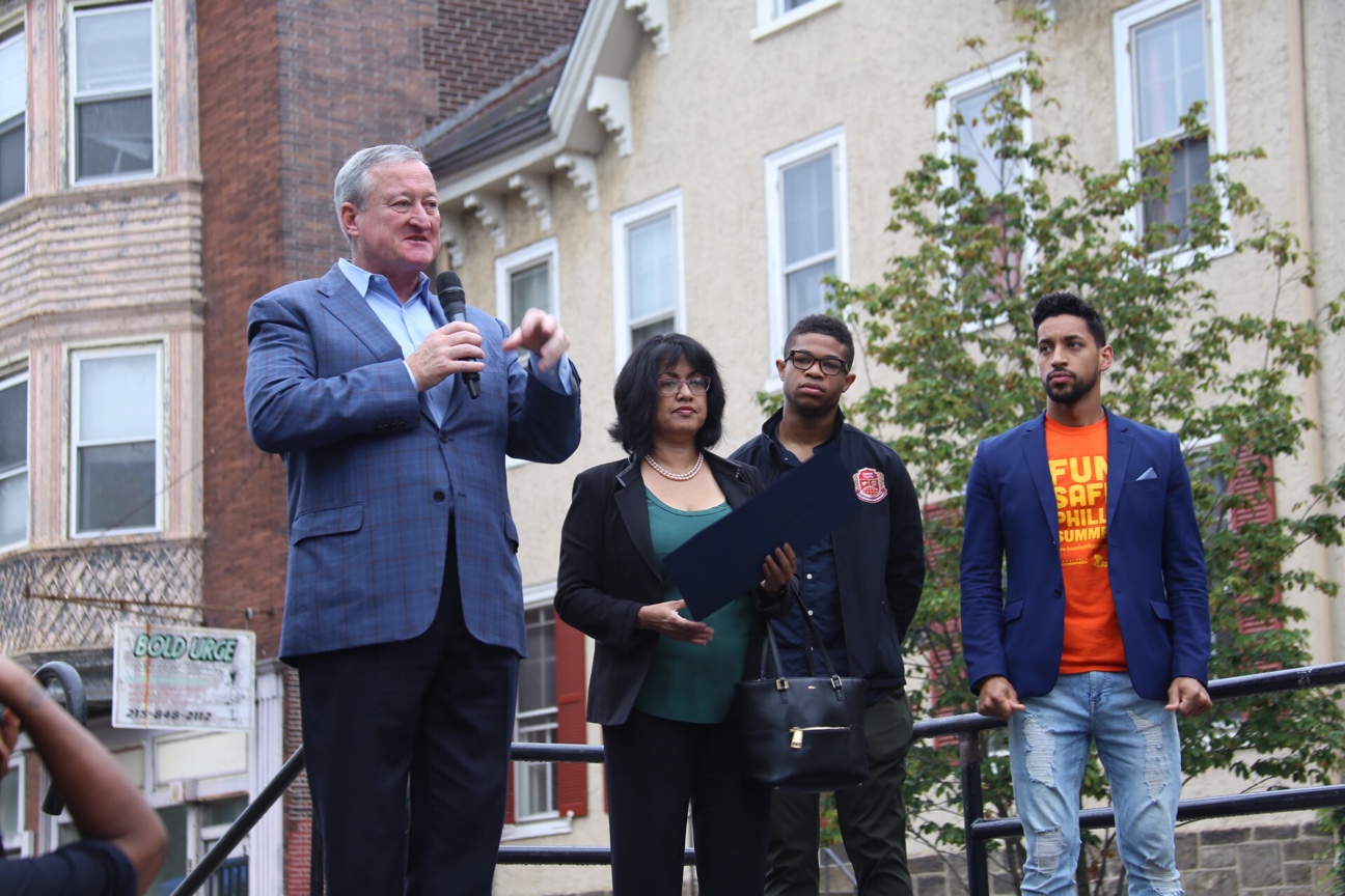 The Mayor is standing speaking with a microphone on a stage with colonial houses behind him and City workers alongside him looking on.