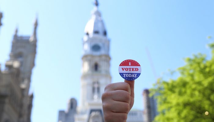 person holding up their thump with an i voted sticker in front of city hall