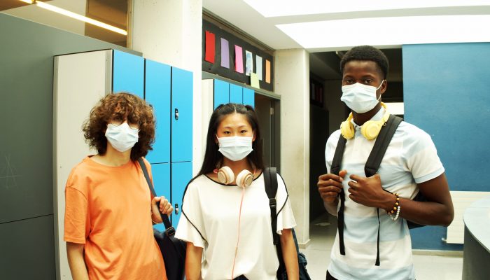 三个学生背着书包、戴着口罩，并肩站在走廊上。