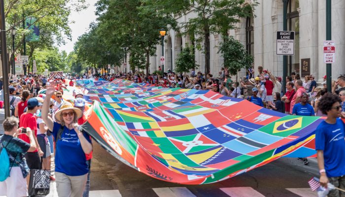La gente camina por una calle de Filadelfia sosteniendo una gigantesca bandera de colores que presenta imágenes más pequeñas de banderas de diferentes países del mundo. Una multitud de personas se alinean a ambos lados de la calle.