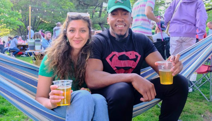 A man and a woman sit on a hammock drinking beer in the park.