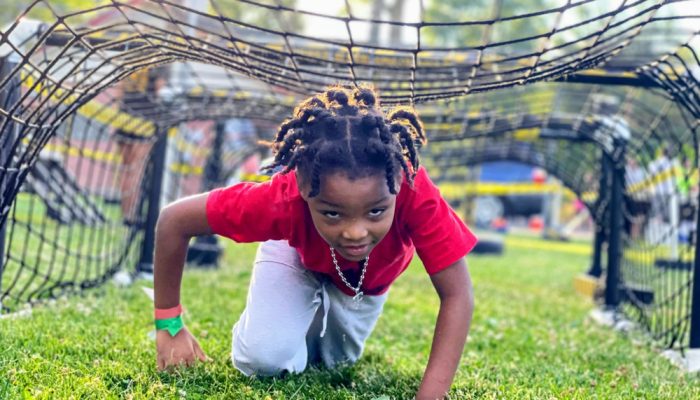 An African-American youth crawls under a large cargo net outside. It is part of a child's obstacle course.
