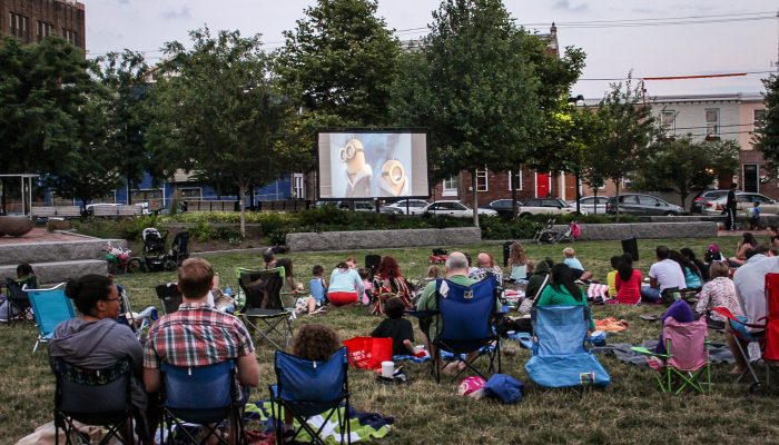 People watch a movie outdoors in the park on a large screen. They are sitting in chairs and blankets.