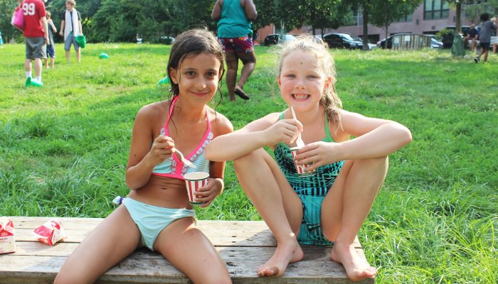 Two little girls eat Italian water ice in the park. The girls are wearing bathing suits and sitting on a bench in a Philadelphia park.