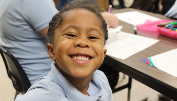 An African-American girl smiles at a Philadelphia Parks & Recreation after school program. She is wearing a light blue polo shirt and sitting at a table.