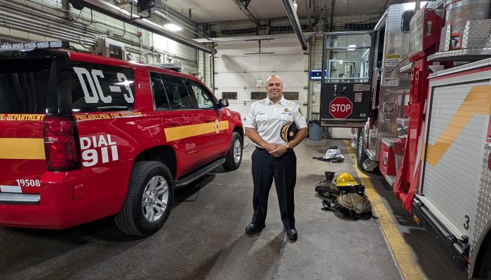 A bald Latino man stands between two Philly Fire Department Vehicles. He's wearing a white shirt, and holding a hat. On the right is a firetruck and on the left is an SUV that says DC3, which means Deputy Chief.