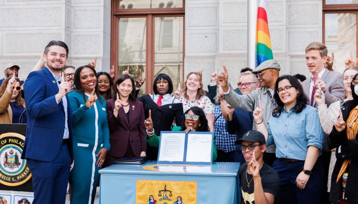 Mayor Cherelle L. Parker sitting behind a desk, holding up a folder, joined by members of her Administration