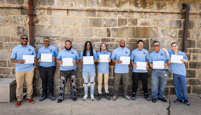 Ten masonry academy graduates in blue shirts holding diplomas in front of a stone wall.