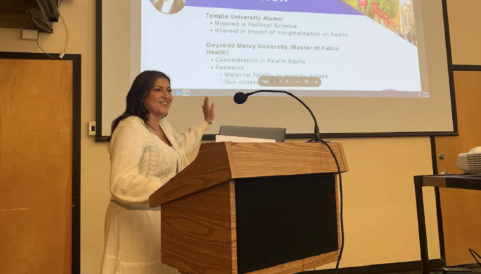 A VISTA member presents at a podium, with a projected slide about her education and research interests in public health and health equity behind her.