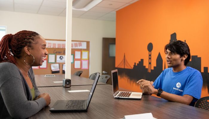 Two individuals seated at a table across from one another, each using a laptop, engaged in a collaborative work session.